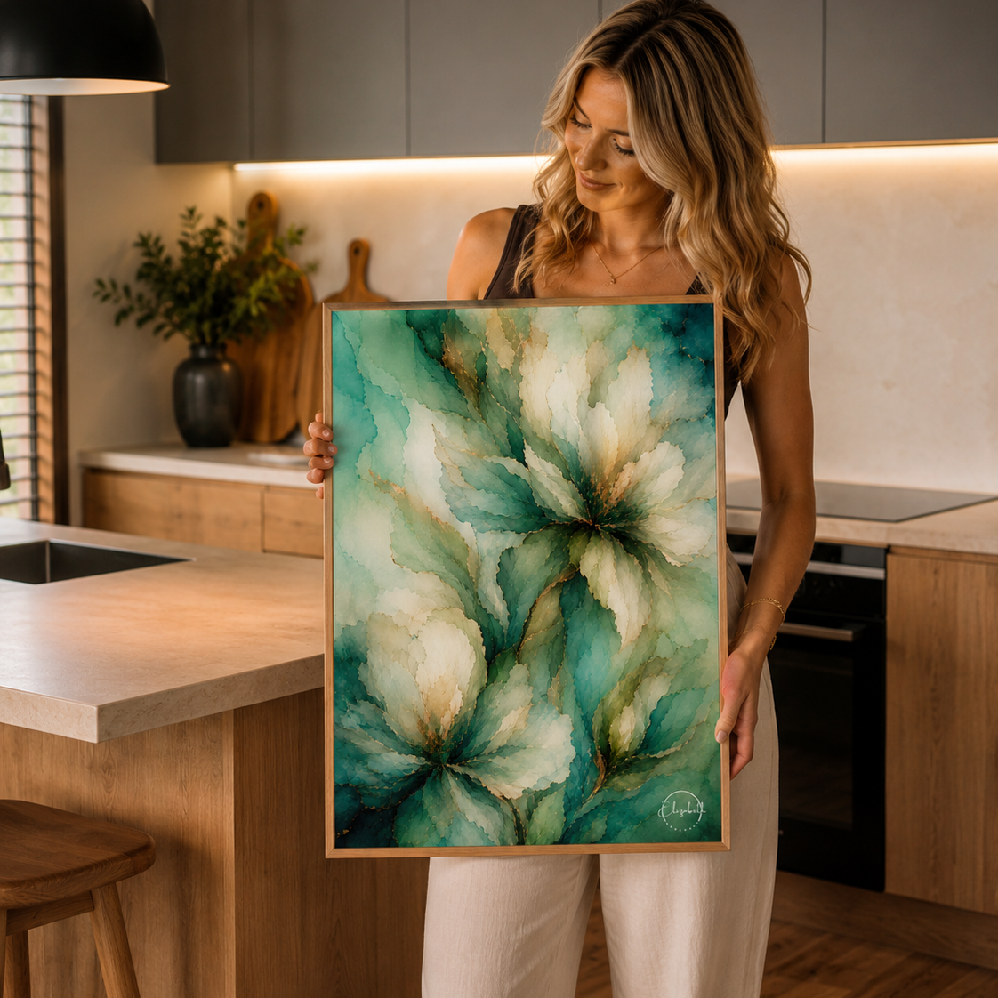 A woman stands in a modern kitchen holding a framed abstract floral painting with teal, green, and cream tones.