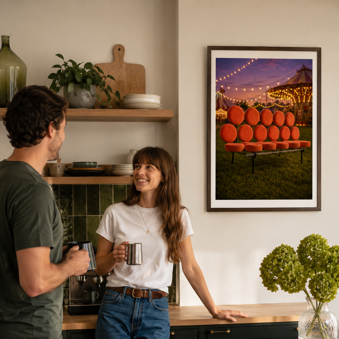 A framed art print hangs on a warm beige wall in a cozy kitchen setting, showing a colorful sunset scene with an orange marshmellow sofa. A smiling couple stands in the kitchen, holding mugs and engaging in conversation, creating a warm, inviting lifestyle atmosphere.