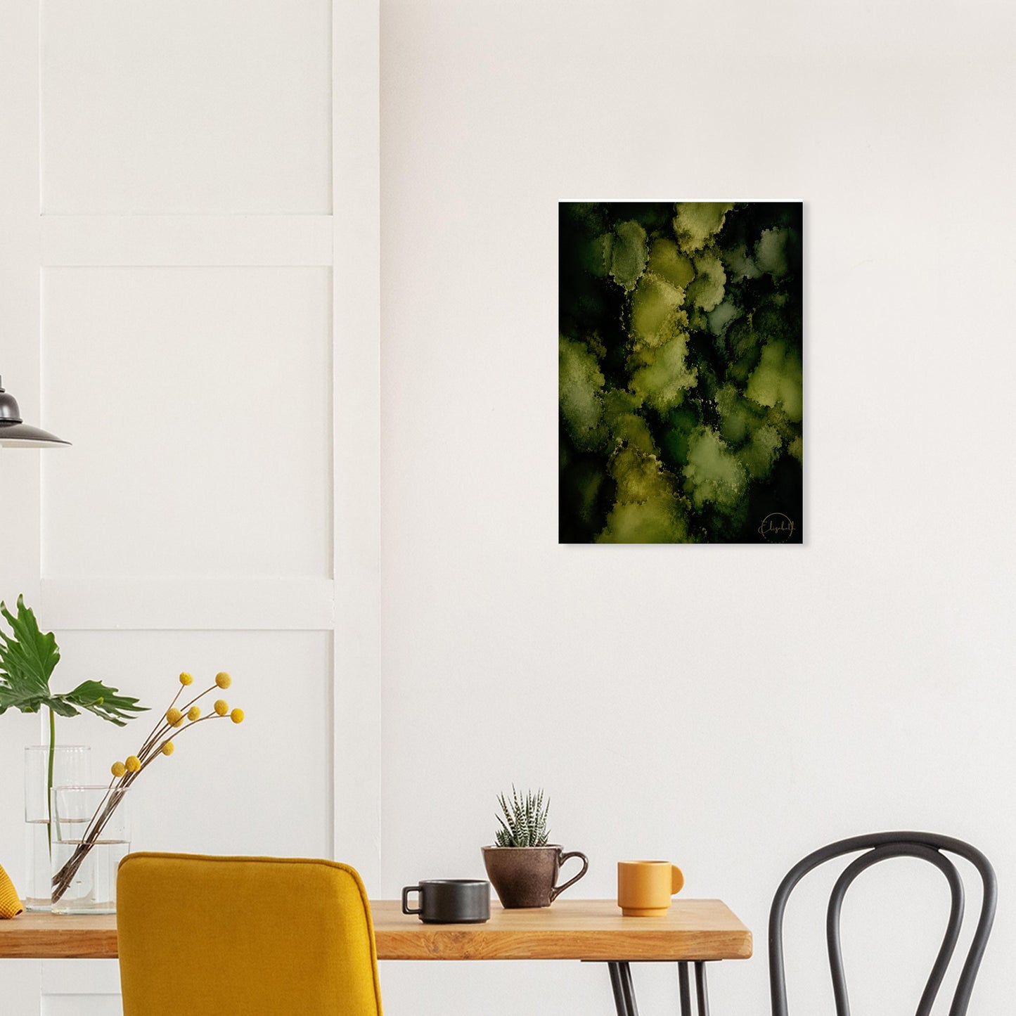 A vertical abstract art print featuring green and yellow-green textured circles on a black background, displayed on a white wall above a dining table with chairs and mugs.