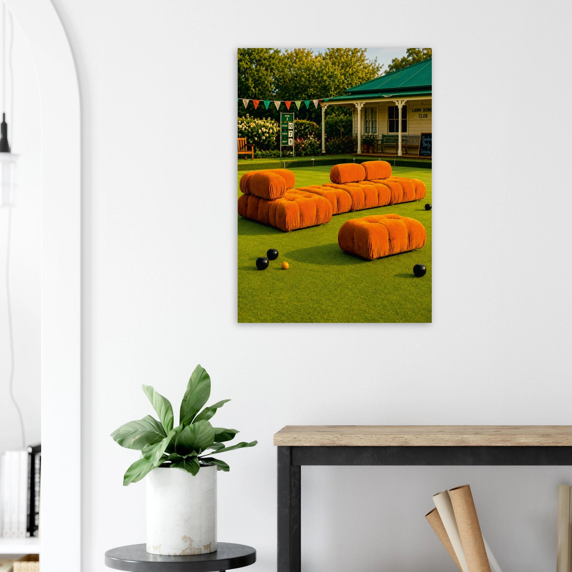 A vertical wall art print depicting an outdoor patio scene with bright orange modular seating on a green lawn, displayed on a white wall above a console table with a potted plant.