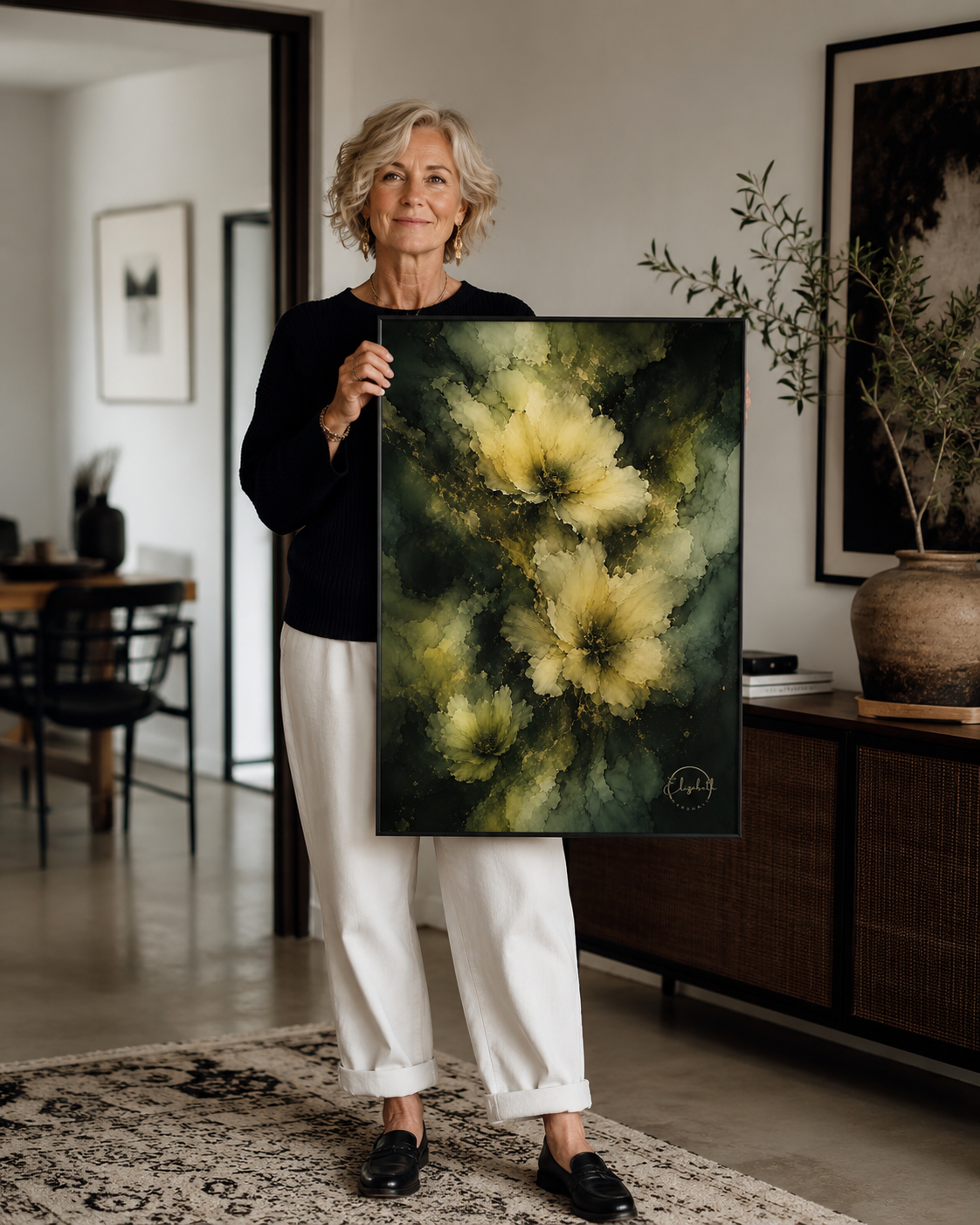 An older woman with short gray hair stands in a modern living room holding a large vertical canvas painting. The painting features abstract yellow and cream floral shapes against a dark green and black textured background.