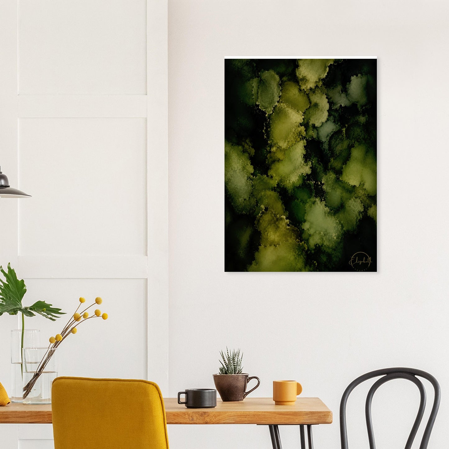 An abstract wall art print featuring textured green and yellow-green organic shapes on a black background, displayed on a white wall above a dining table with chairs and mugs.