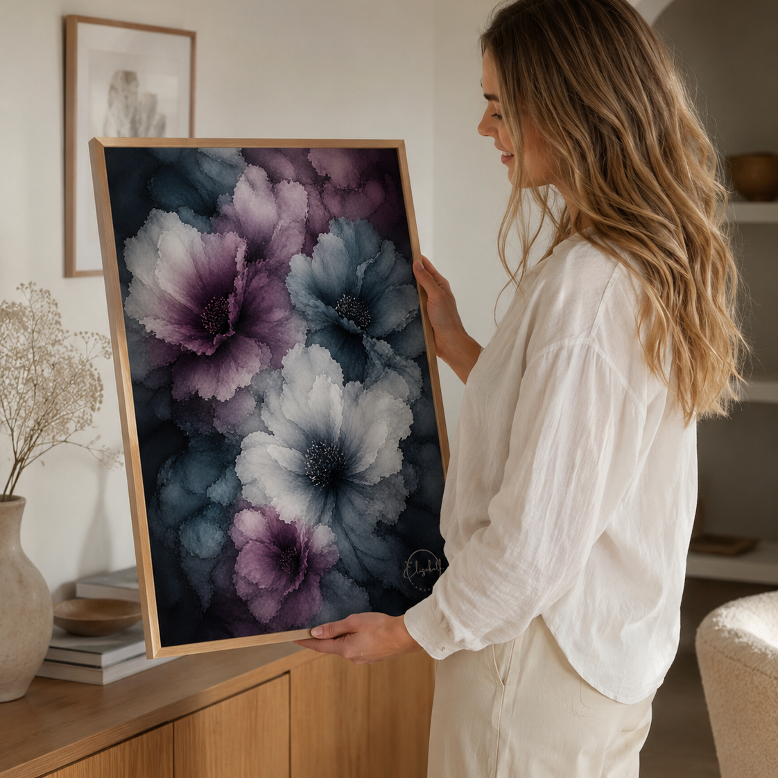 A woman in a white outfit holds up a framed abstract floral print featuring large purple, blue, and white flowers against a dark background, set in a warm, modern interior room.