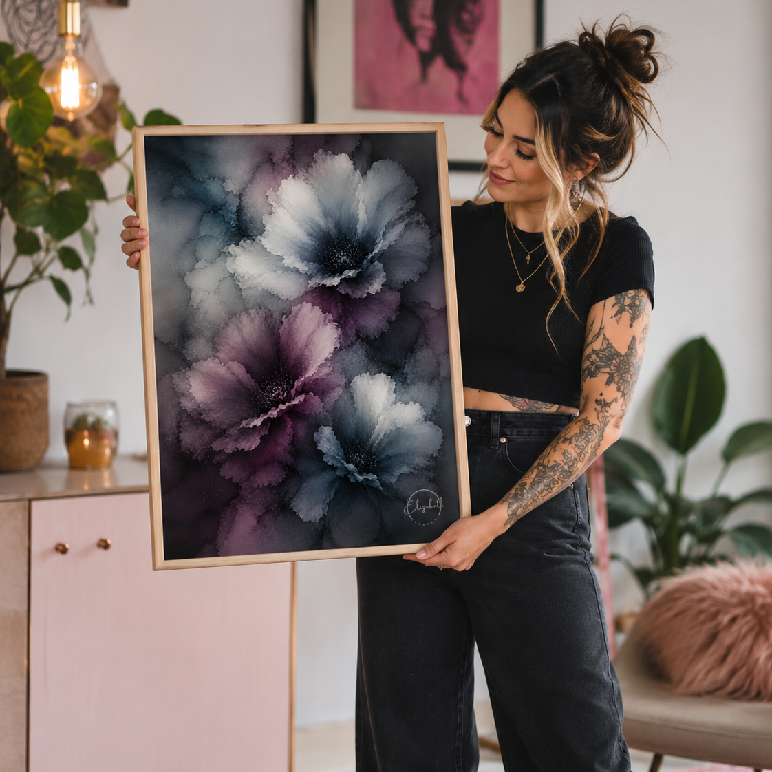 A woman with arm tattoos holds up a framed abstract floral art print featuring large purple, blue, and gray flowers. She is standing in a modern, stylishly decorated room with a pink cabinet, plants, and warm lighting.