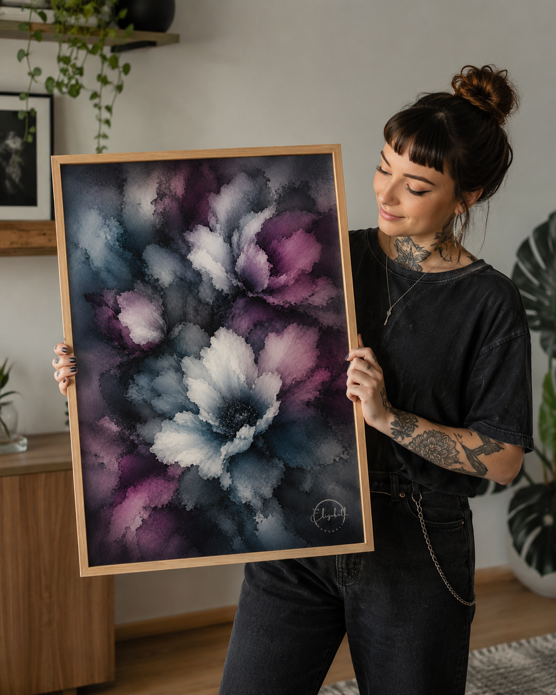 A woman with tattoos holds up a large framed abstract floral art print featuring purple, white, and blue watercolor-style petals. The print has a light natural wood frame.
