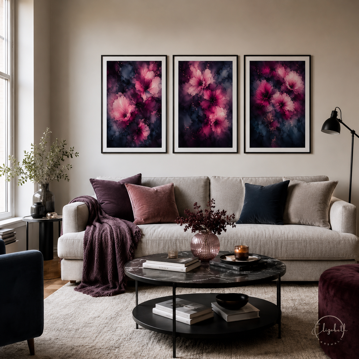 A styled living room with a light beige sofa, dark accent pillows, and a round coffee table, featuring three matching framed abstract floral wall art prints in pink, purple, and black above the couch.
