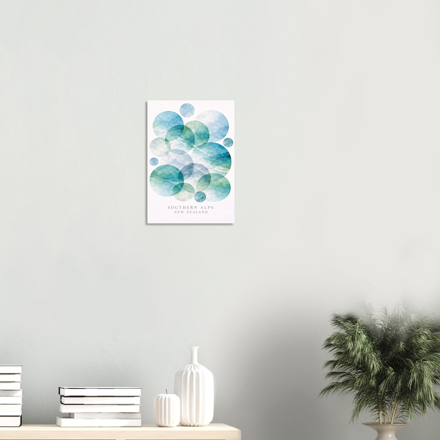 A vertical wall art print featuring a watercolor design of overlapping blue and green circles on a white background, styled on a light gray wall above a shelf with books, a vase, and a potted plant.