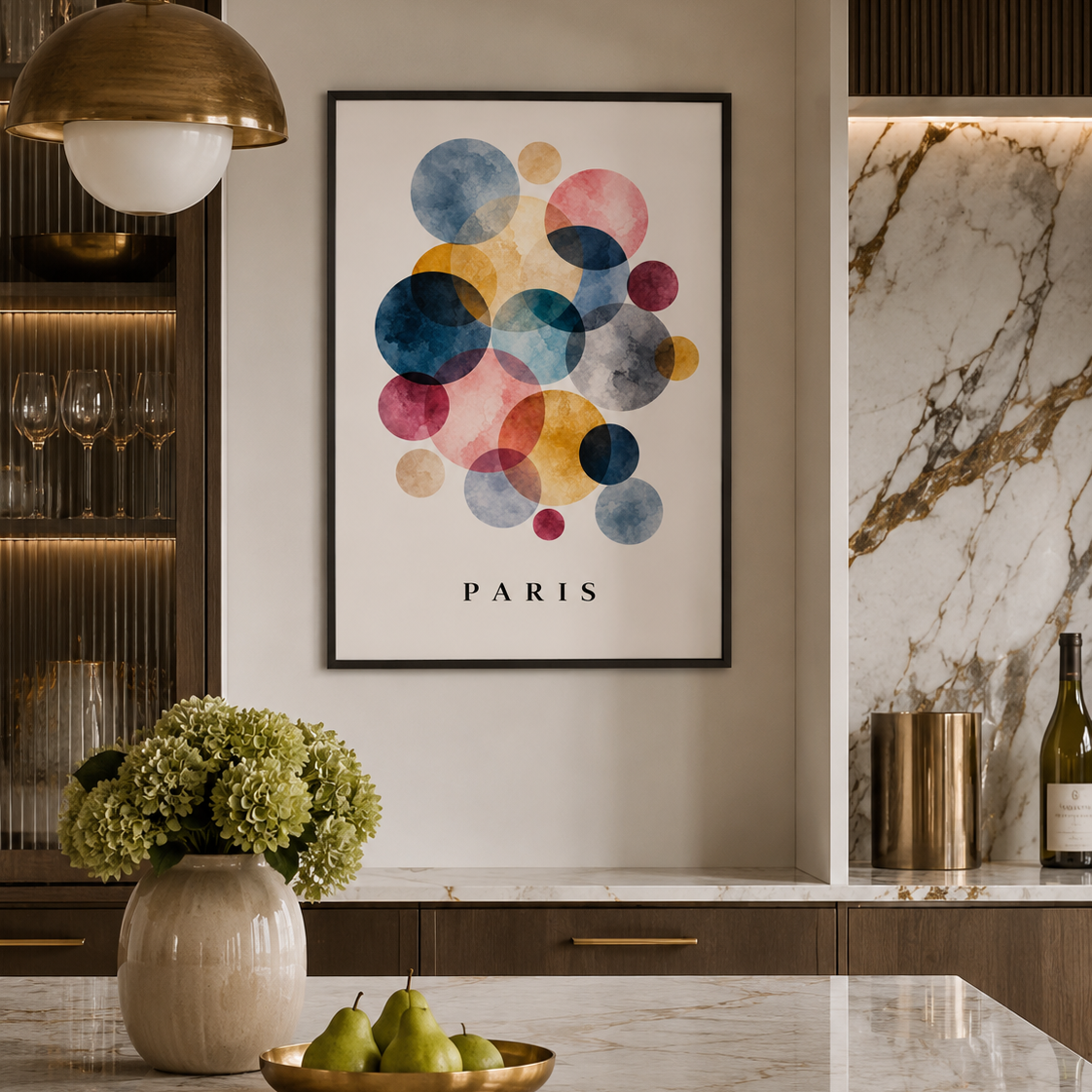 A framed wall art print featuring colorful watercolor circles above the word PARIS, displayed in a modern kitchen setting with marble surfaces, a hanging brass pendant light, and green pears on a counter.
