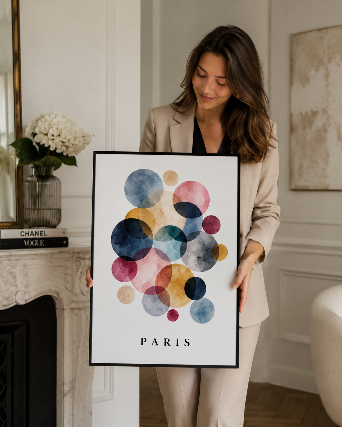 A woman in a beige blazer holds a framed art print featuring colorful watercolor circles on a white background with the word 'PARIS' at the bottom, set in a bright, elegant room.
