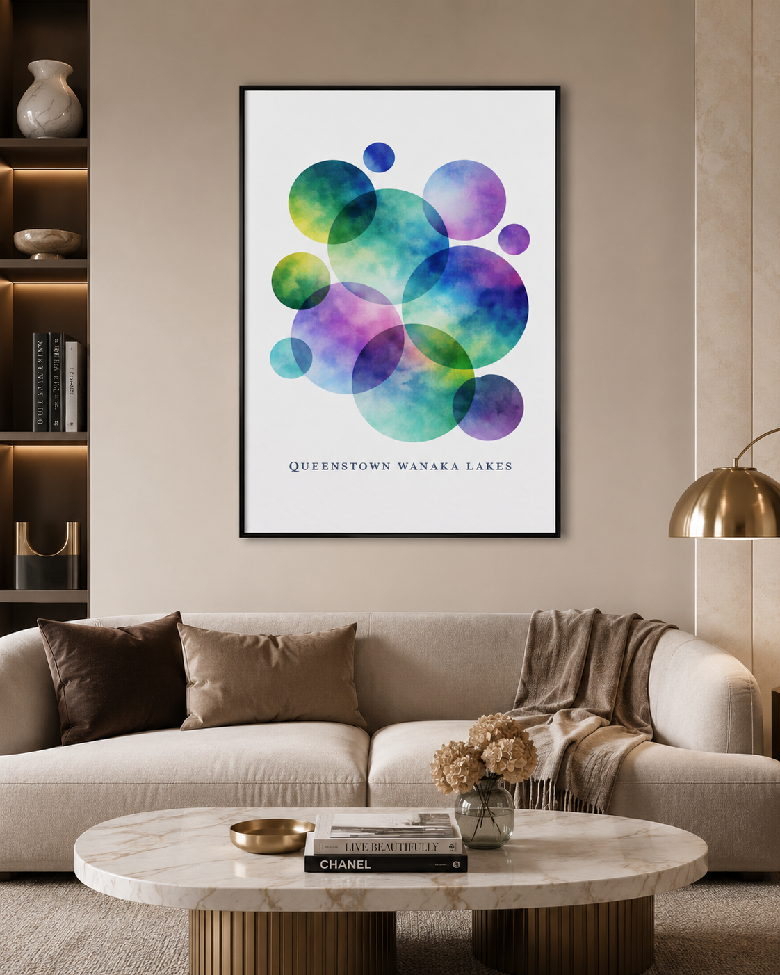 A framed wall art print featuring overlapping watercolor circles in green, blue, purple, and teal above the text 'QUEENSTOWN WANAKA LAKES', displayed above a beige sofa in a modern living room setting.