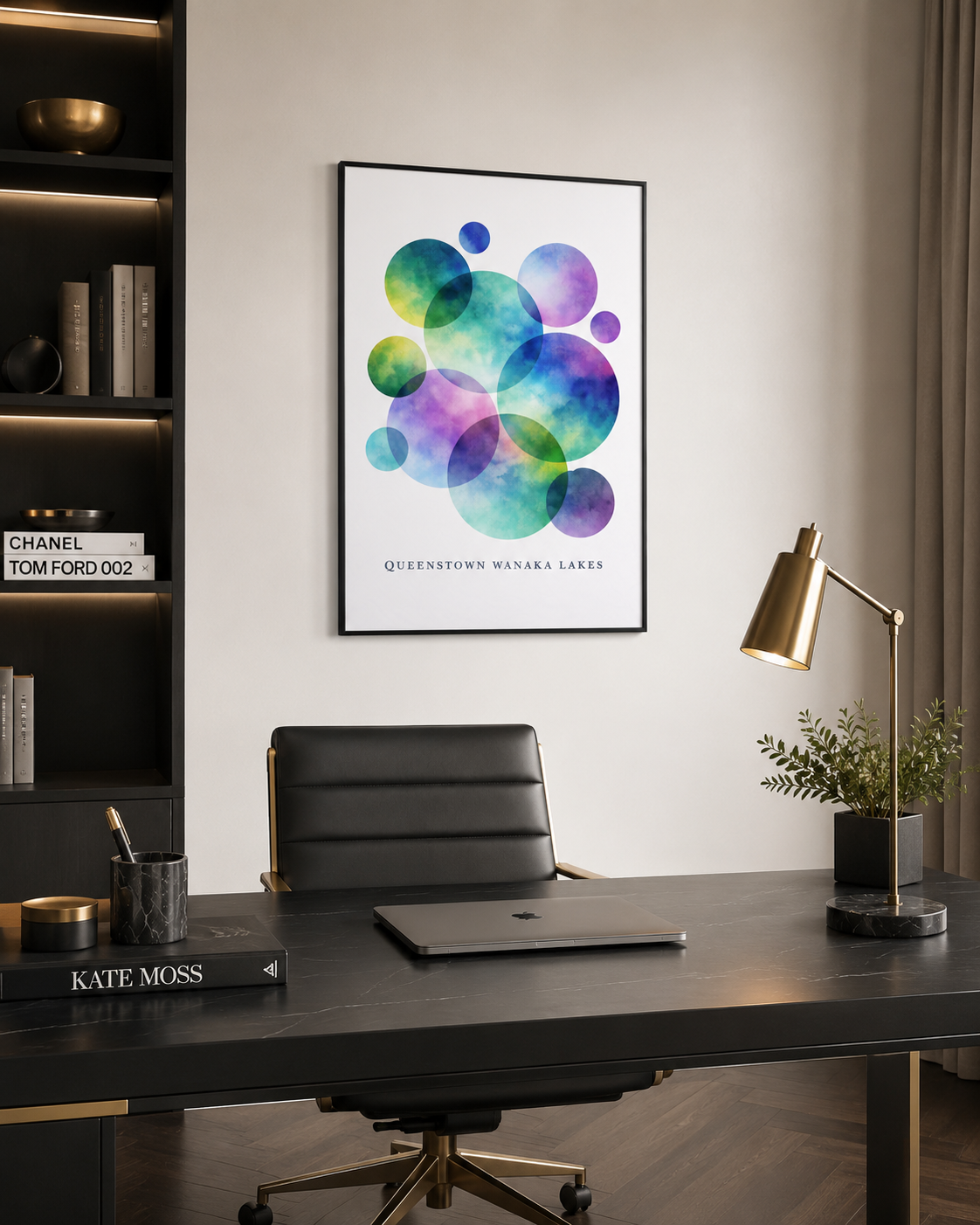 A framed art print featuring colorful watercolor circles in shades of blue, green, purple, and teal, with the text 'QUEENSTOWN WISCONSIN LAKES' at the bottom. The print is displayed on a light gray wall above a modern black desk with a laptop, office chair, and a gold desk lamp.