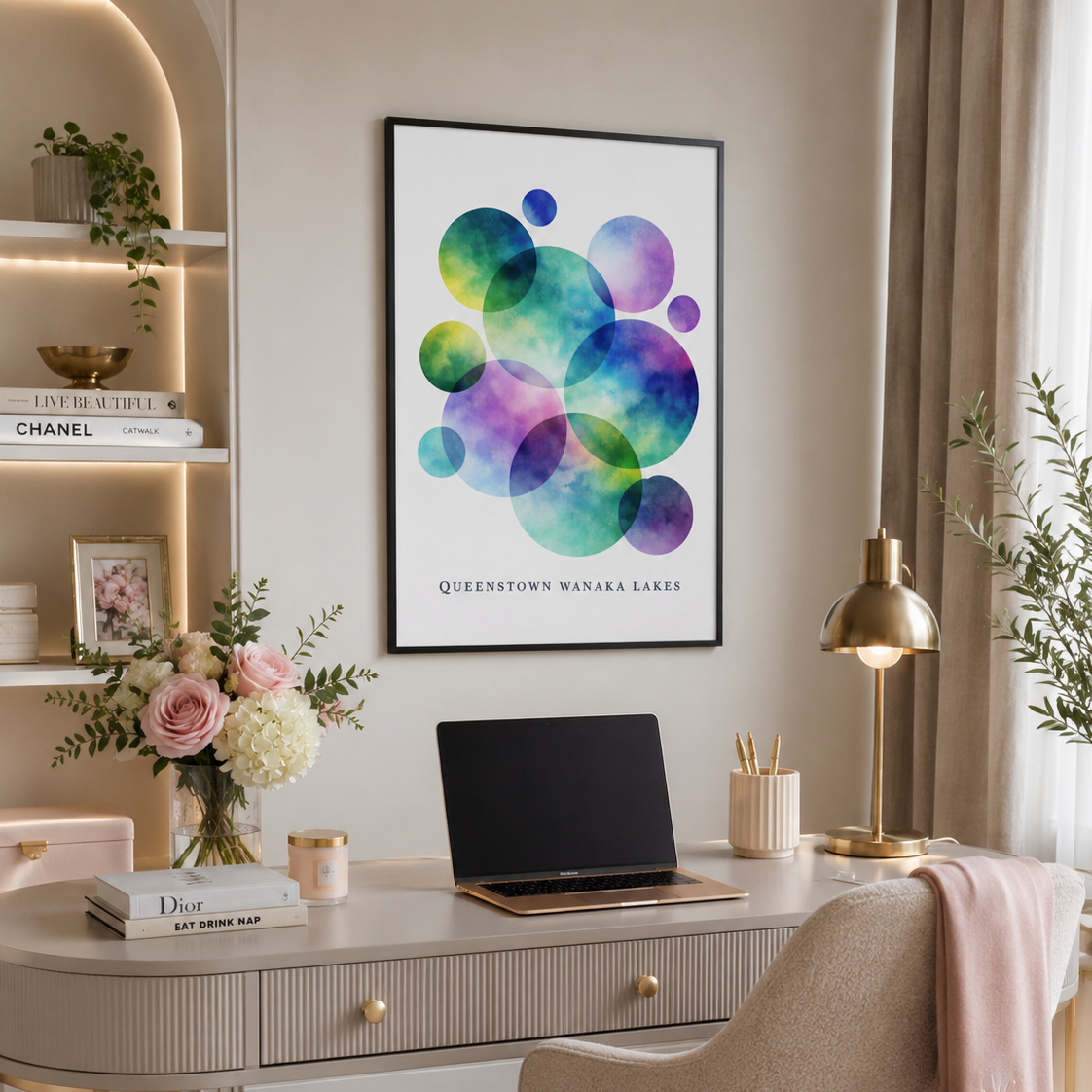 A framed wall art print featuring colorful overlapping watercolor circles in green, blue, purple, and teal, with the text 'QUEENSTOWN WANAKA LAKES at the bottom. The print is displayed in a styled home office setting above a beige desk with a laptop, lamp, and chair.
