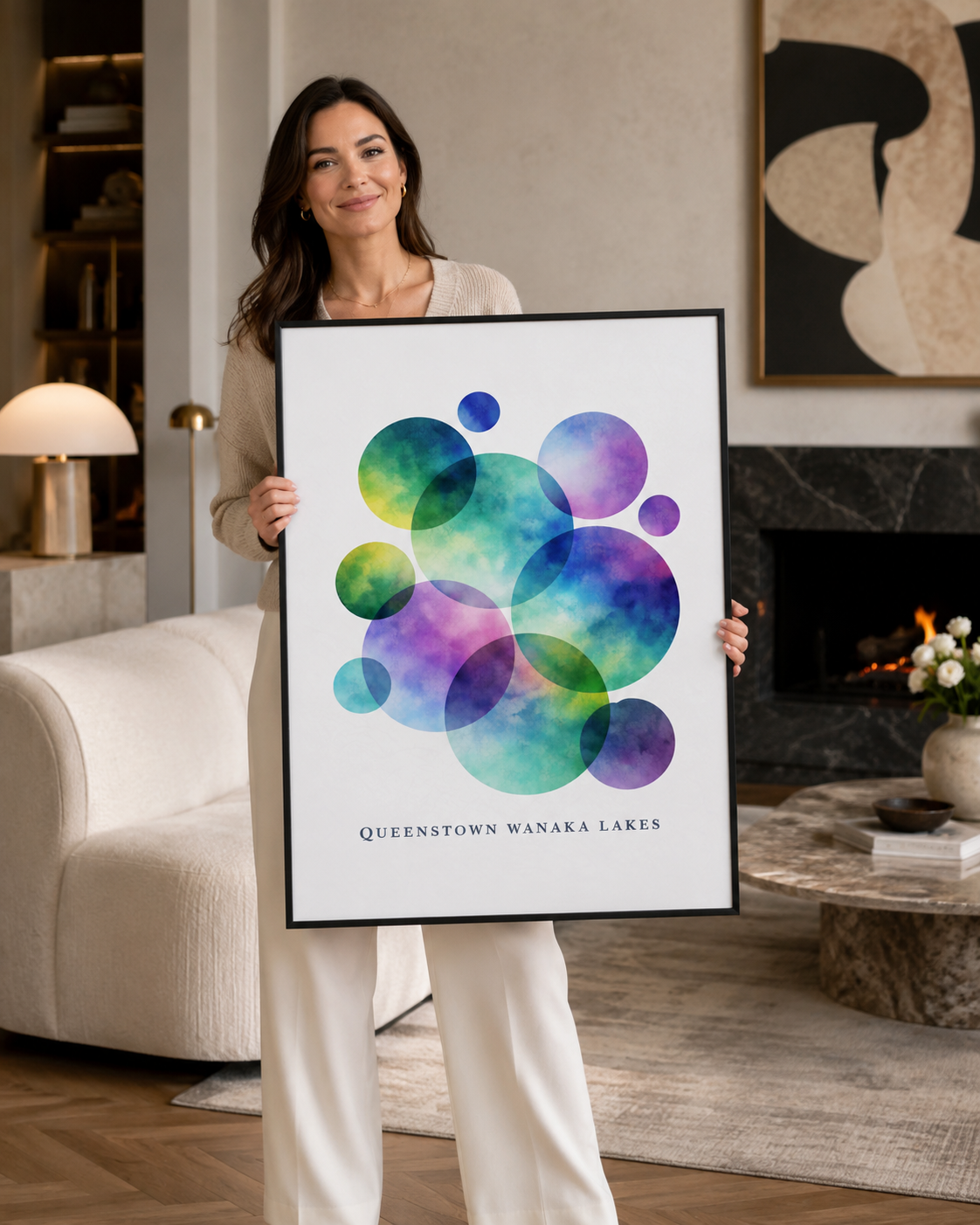 A woman stands in a modern living room holding a large framed art print. The print features overlapping watercolor circles in shades of green, blue, purple, and teal on a white background, with the text 'QUEENSTOWN WANAKA LAKES' printed at the bottom.