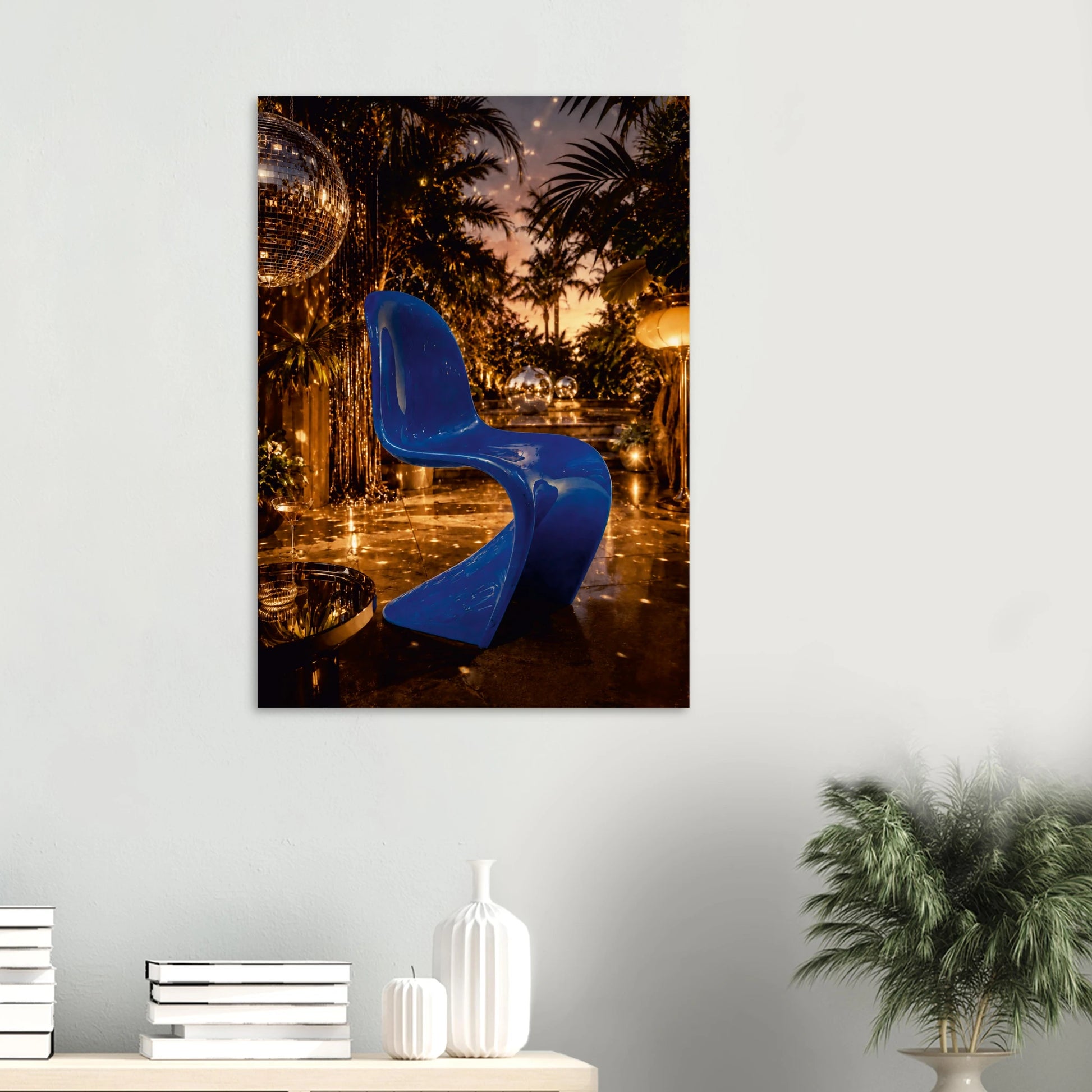 A vertical wall art print depicting a glossy blue modern chair in a tropical nighttime setting with palm trees and warm lights, displayed on a light gray wall above a console table with books and a plant.