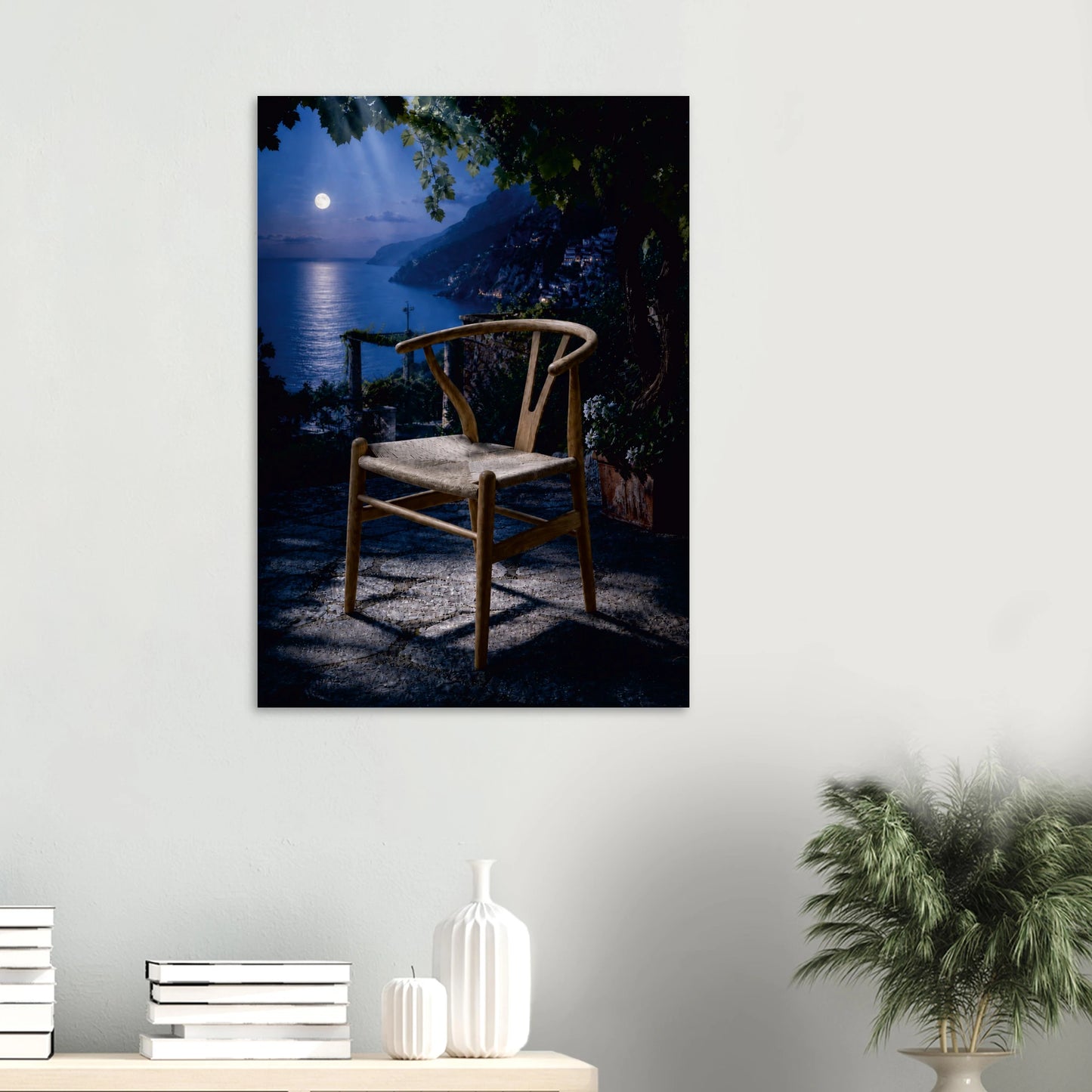 A framed wall art print depicting a wooden chair in front of a night ocean scene with moonlight, displayed on a light gray wall above a console table with books and decor.
