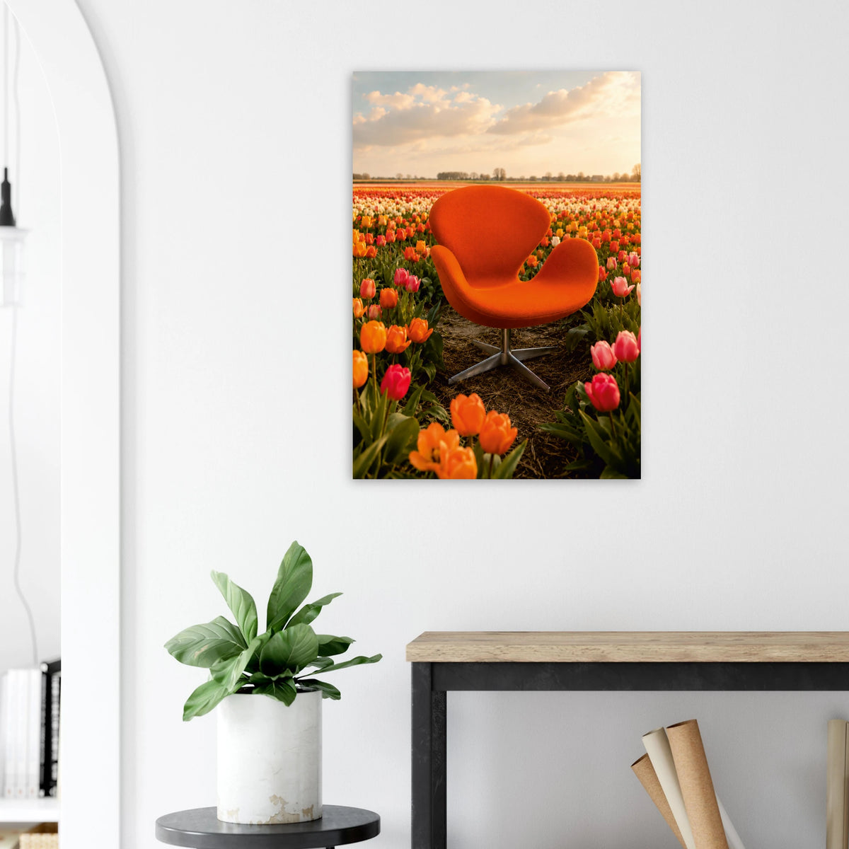 A vertical canvas wall art print depicting an orange chair in a tulip field at sunset, displayed on a white wall above a console table with a potted plant.