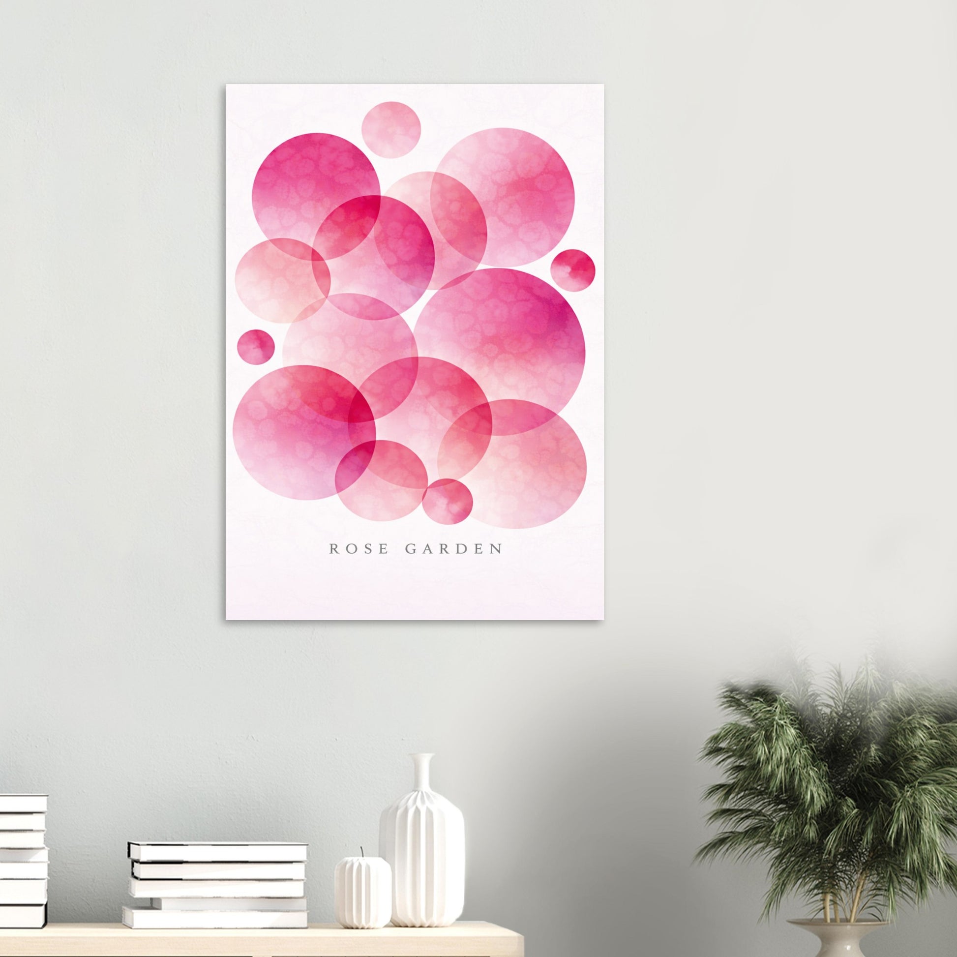 A vertical wall art print featuring overlapping pink watercolor circles on a white background with the text 'ROSE GARDEN' near the bottom, displayed on a light gray wall above a console table with books, a vase, and a potted plant.