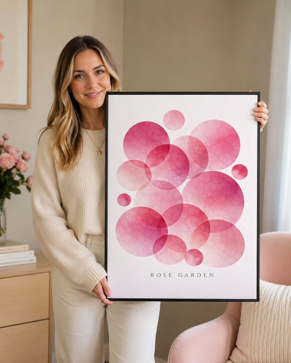 A woman in a cream ribbed sweater and matching pants holds up a framed art print featuring overlapping pink watercolor circles with the text 'ROSE GARDEN' at the bottom. The setting is a cozy, modern room with a pink armchair and a vase of pink roses on a light wood side table.