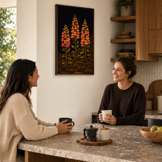 A framed vertical wall art print featuring three stylized orange and red flowers on a dark background hangs on a beige wall in a modern kitchen. Two women are seated at a marble kitchen island, holding mugs and engaging in conversation.