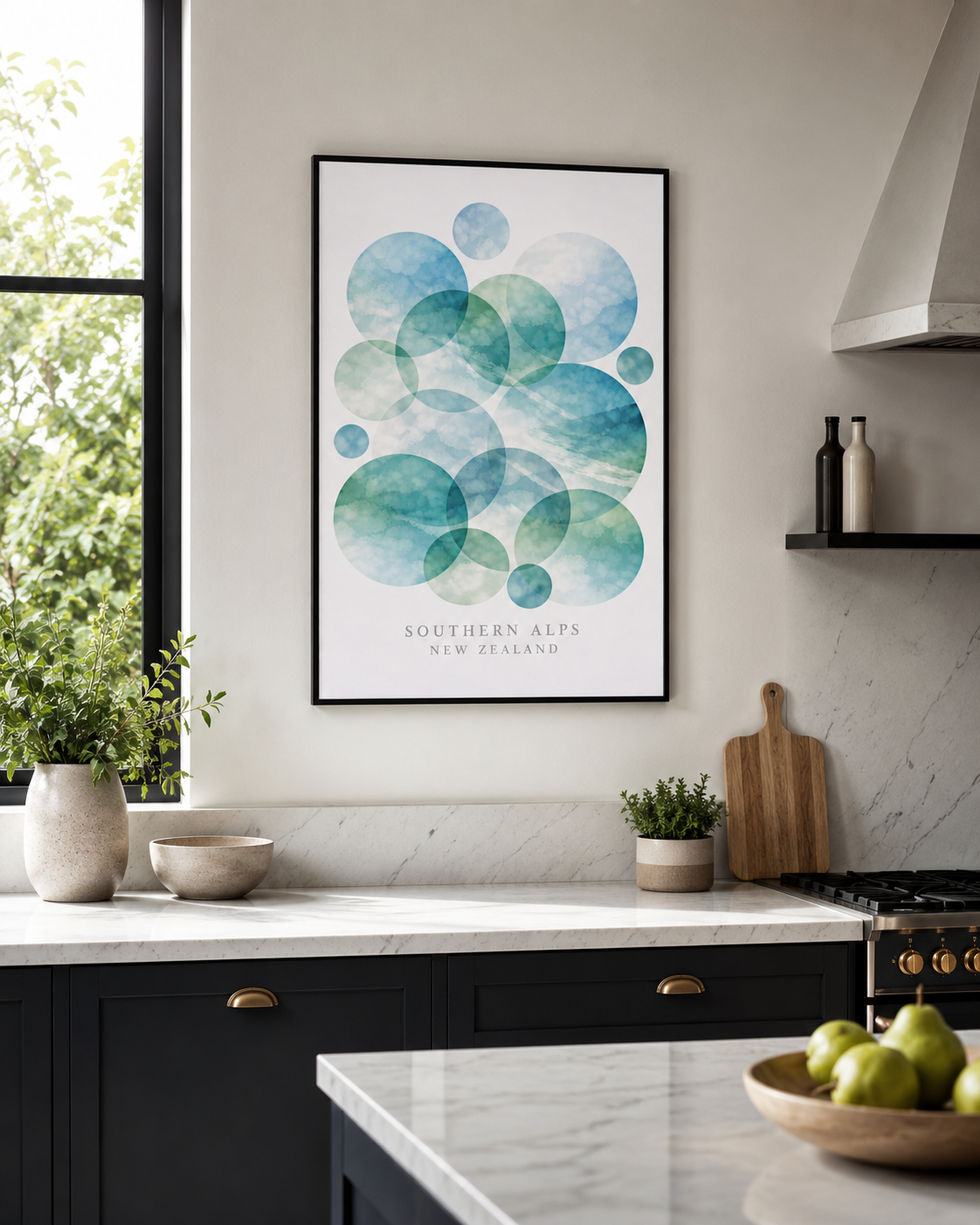 A framed art print featuring overlapping watercolor circles in shades of teal, blue, and green, with the text 'SOUTHEASTERN ALPS NEW ZEALAND' at the bottom. The print is displayed on a light gray wall in a modern kitchen setting with dark cabinets, marble countertops, and green pears on a table in the foreground.
