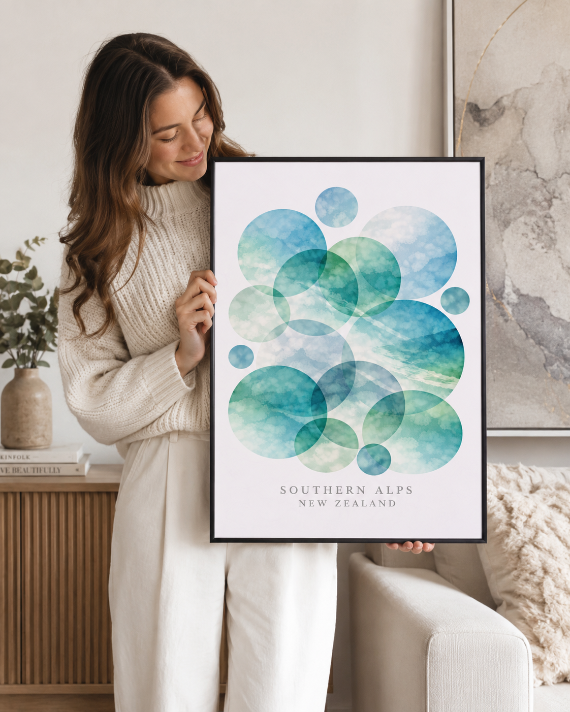 A woman in a cream knit sweater holds up a framed art print featuring watercolor-style overlapping circles in shades of teal, blue, and green, with the text 'SOUTHERN ALPS NEW ZEALAND' at the bottom. The setting is a bright, modern living room with a beige sofa and neutral decor.