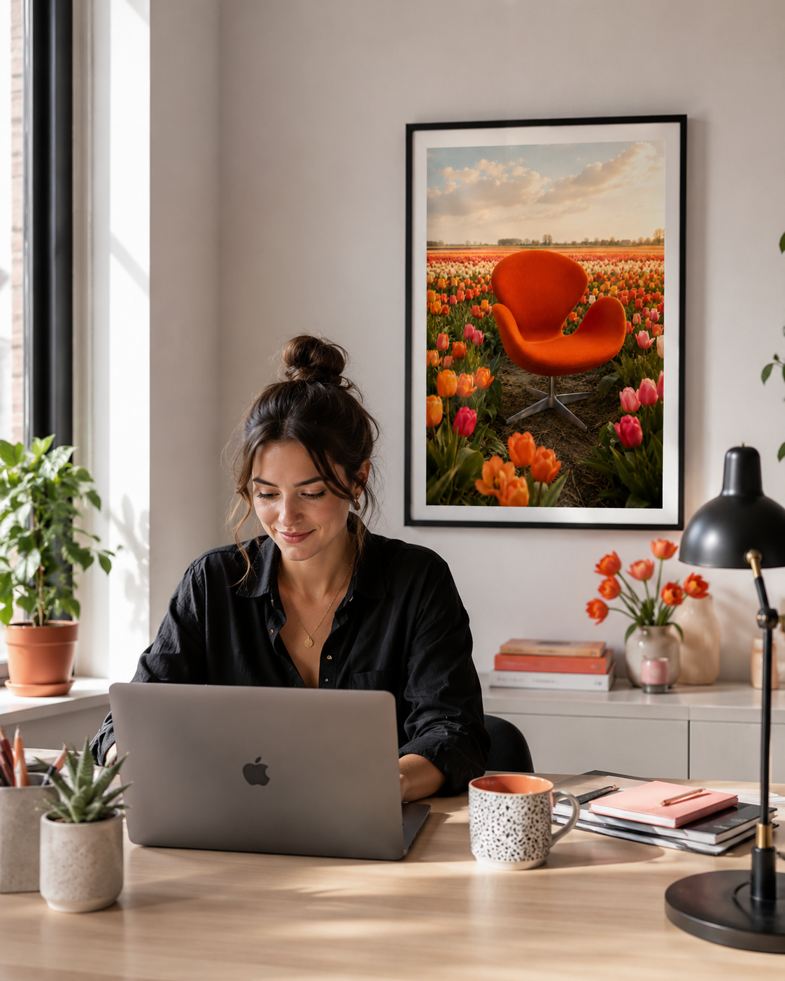 A woman sits at a wooden desk using a laptop in a bright home office, with a framed art print of an orange chair in a tulip field hanging on the wall behind her.
