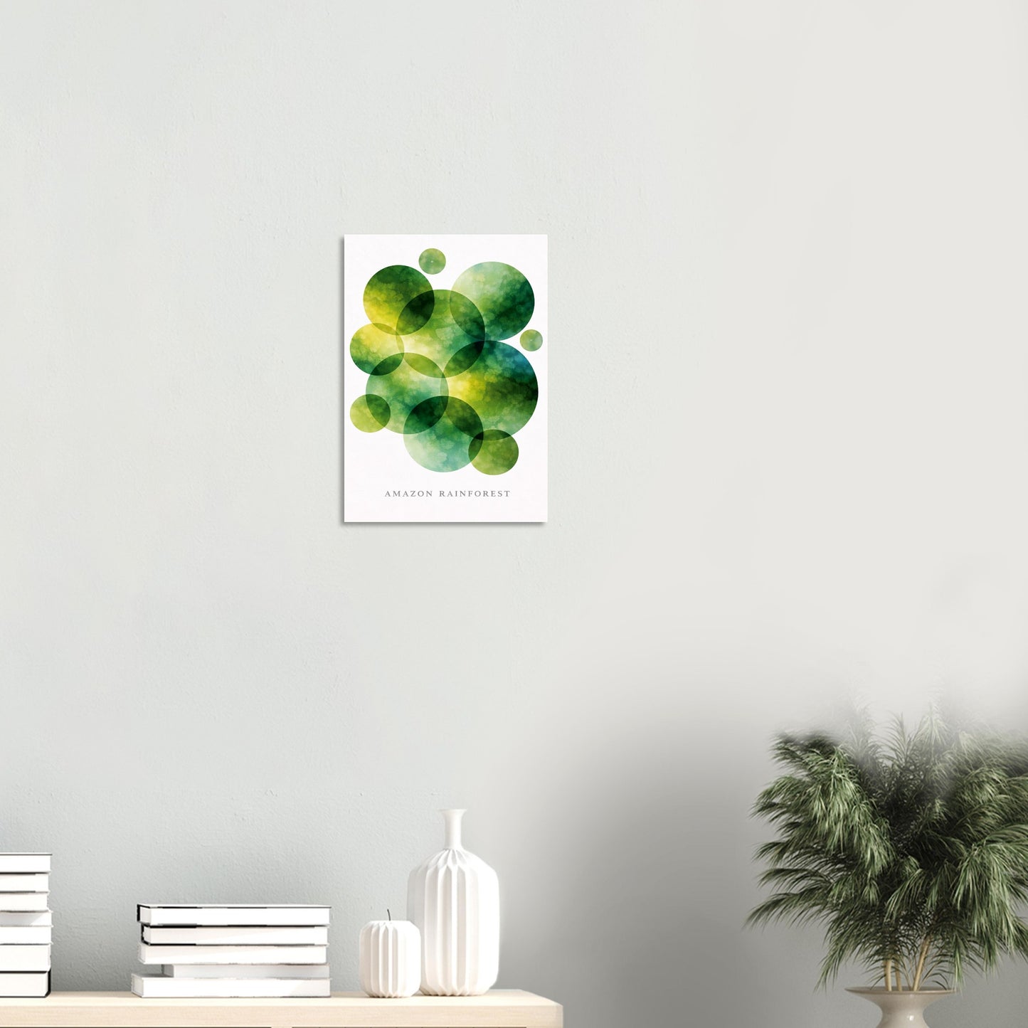 A framed wall art print featuring overlapping green and yellow watercolor circles on a white background, hanging on a light gray wall above a shelf with books, a white vase, and a potted plant.