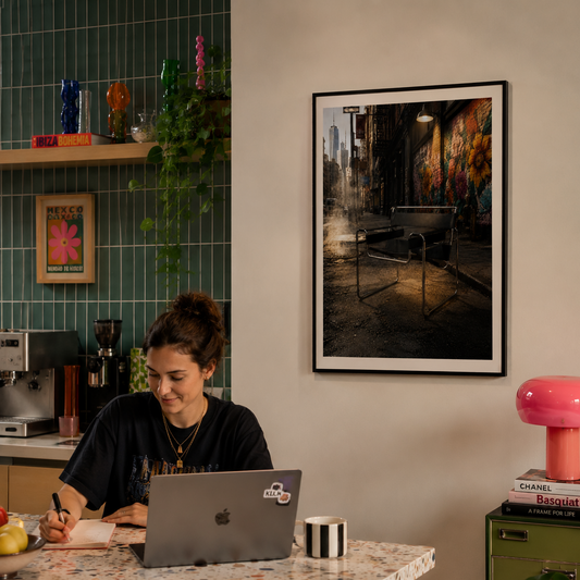 A woman sits at a kitchen island working on a laptop in a modern kitchen, with a framed black-and-white street art photograph hanging on the beige wall above her. The Chair was Looking for Trouble and found exactly the right alley. A Wassily Chair-inspired piece, its angular tubular steel frame and leather slings unmistakable even in the dark, sits alone on rain-slicked cobblestones as a warm street lamp burns overhead. Vivid graffiti murals blaze on the wall behind it, the wet ground reflects the light bel