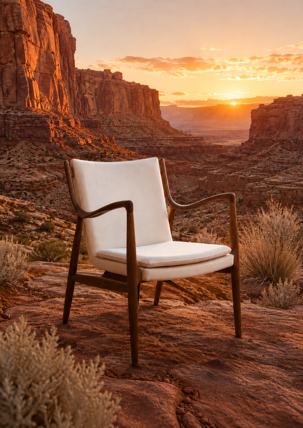A mid-century modern armchair with white cushions and a dark wood frame, set against a dramatic desert canyon landscape at sunset.