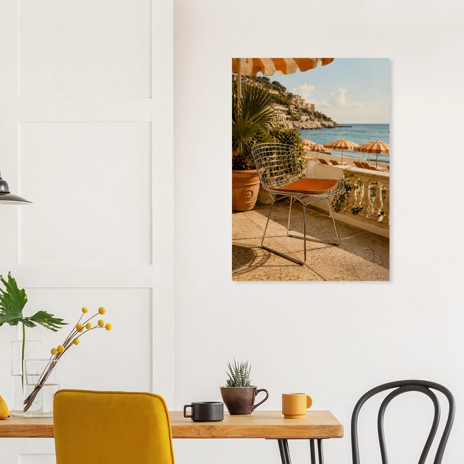 A vertical canvas wall art print depicting a sunny coastal terrace with a wire chair, potted palm, and beach umbrellas, displayed on a white wall above a dining table in a modern room setting.