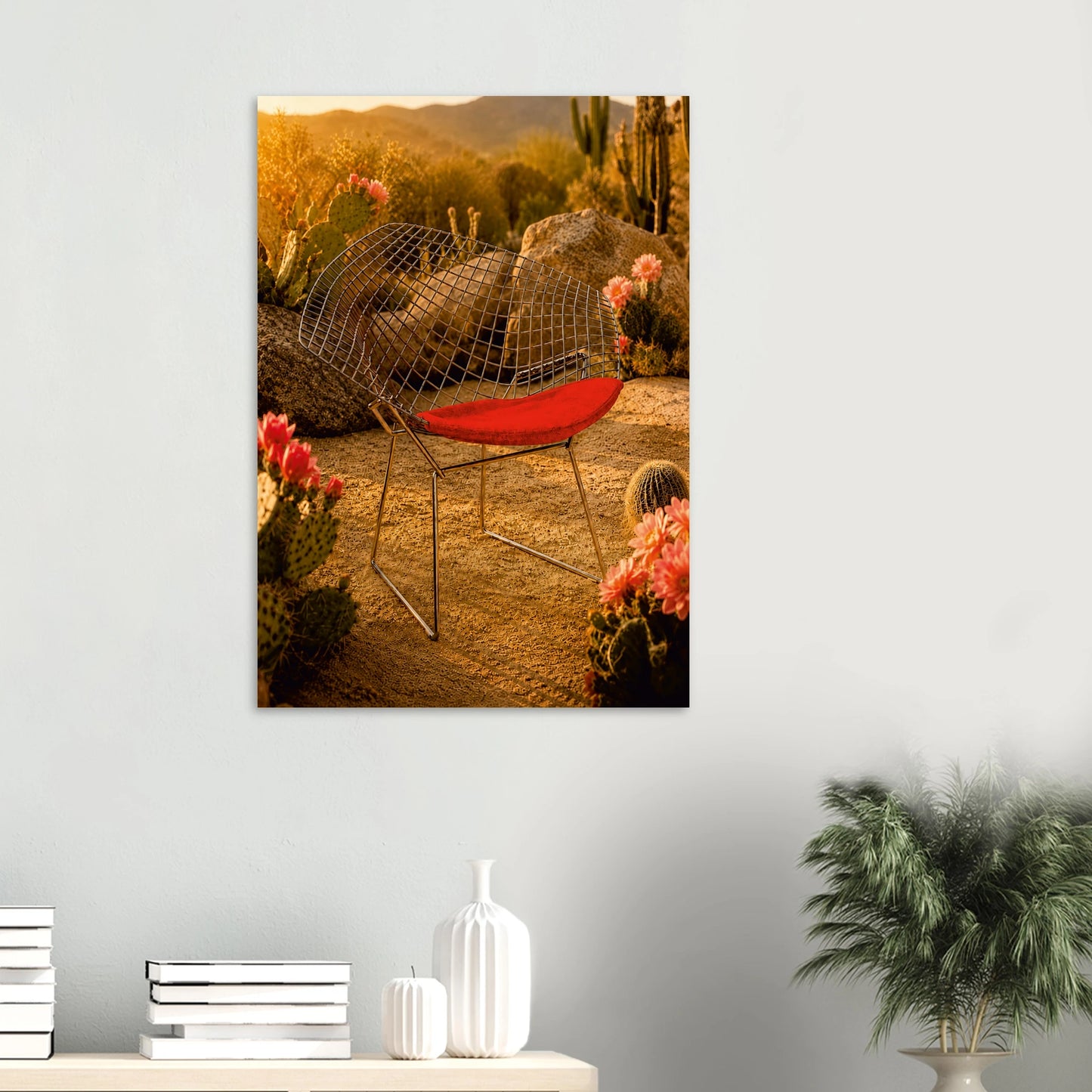 A framed wall art print depicting a desert outdoor scene with a red chair, cacti, rocks, and warm golden light, displayed on a light gray wall above a console table with books and decor.