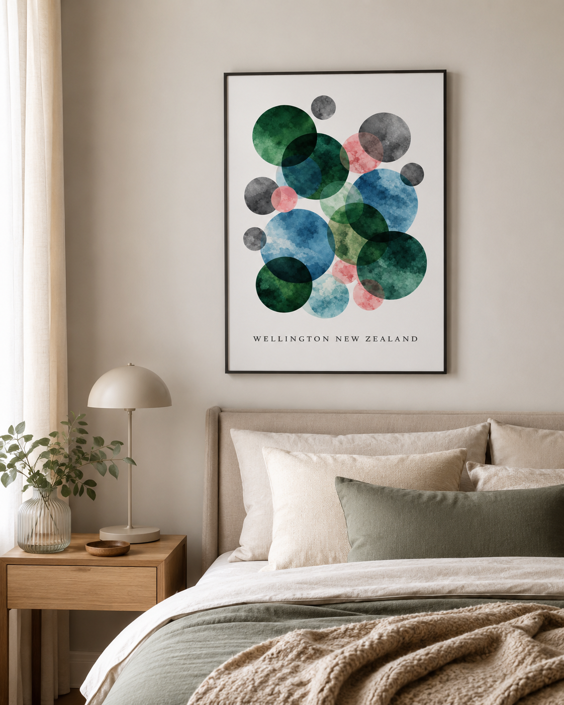 A framed abstract art print featuring overlapping watercolor circles in green, blue, pink, and gray on a white background, with the text 'WELLINGTON NEW ZEALAND' at the bottom. The print is displayed on a beige wall above a bed with neutral-toned bedding and a wooden bedside table with a lamp.