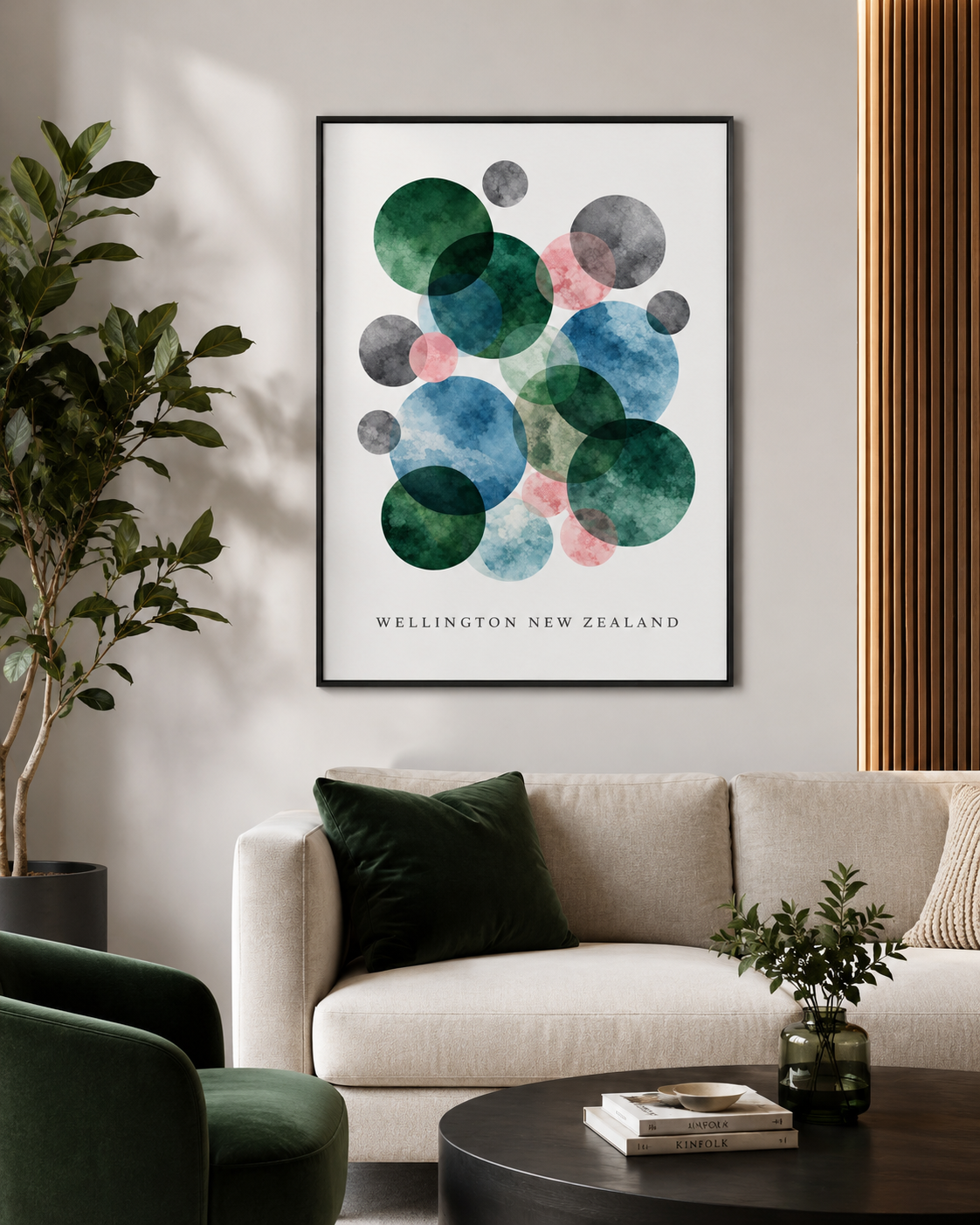 A framed abstract art print featuring overlapping watercolor circles in green, blue, pink, and gray on a white background, with the text 'WELLINGTON NEW ZEALAND' at the bottom. The print is displayed on a light beige wall above a modern beige sofa in a styled living room setting.