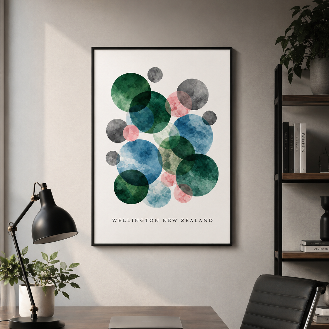 A framed abstract art print featuring overlapping watercolor circles in green, blue, pink, and gray on a white background, with the text 'WELLINGTON NEW ZEALAND' at the bottom. The print is displayed on a light gray wall above a wooden desk with a black lamp, laptop, and chair.