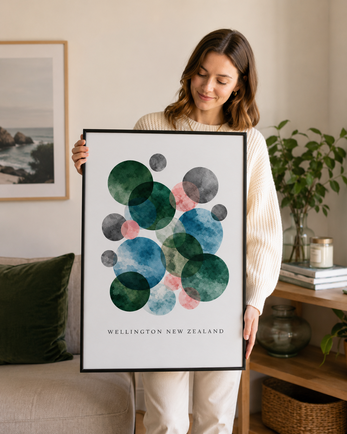 A woman in a cream ribbed sweater holds up a framed art print featuring an abstract watercolor design of overlapping circles in shades of green, blue, pink, and gray, with the text 'WELLINGTON NEW ZEALAND' at the bottom.