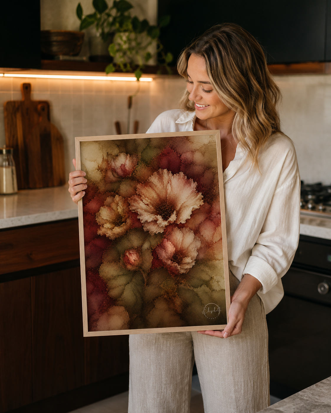 A woman in a kitchen holds up a framed watercolor art print featuring large, textured flowers in shades of dusty pink, rust red, and olive green.