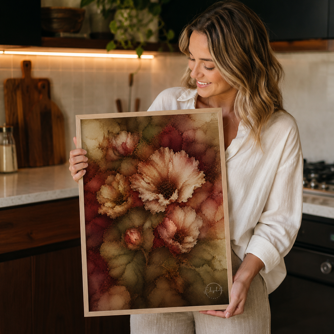A woman in a kitchen holds up a framed watercolor art print featuring large, textured flowers in shades of dusty pink, rust red, and olive green.