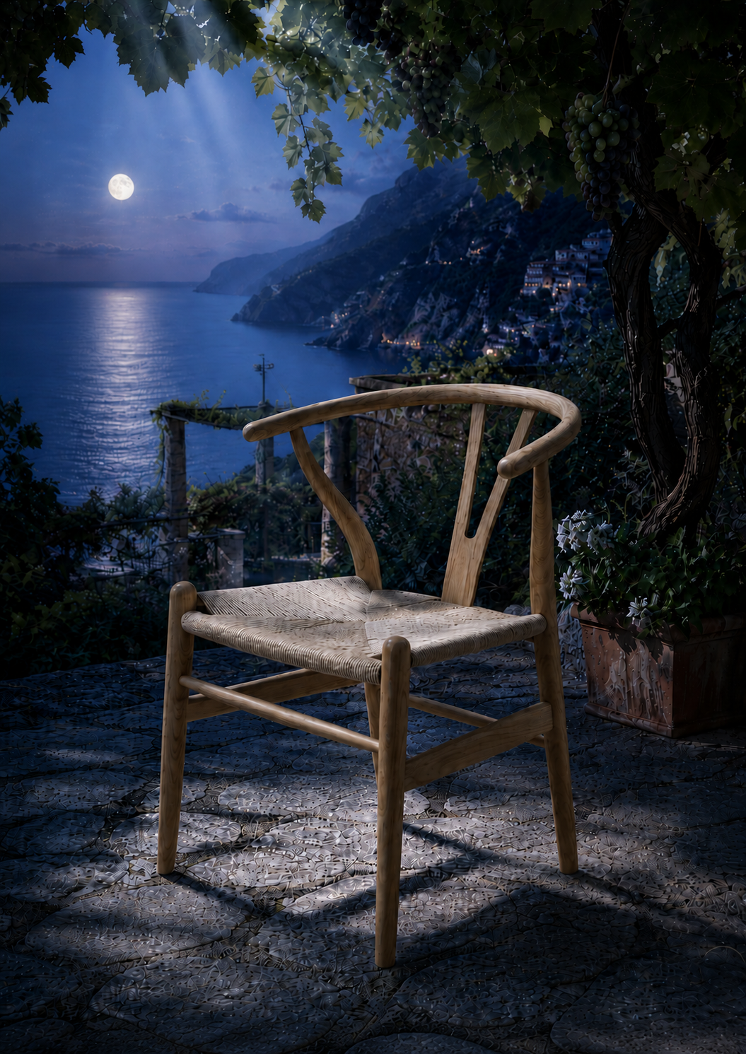 A light natural wood Y-chair with a woven seat sits on a stone patio at night, overlooking a scenic coastal bay with mountains and moonlit sky.
